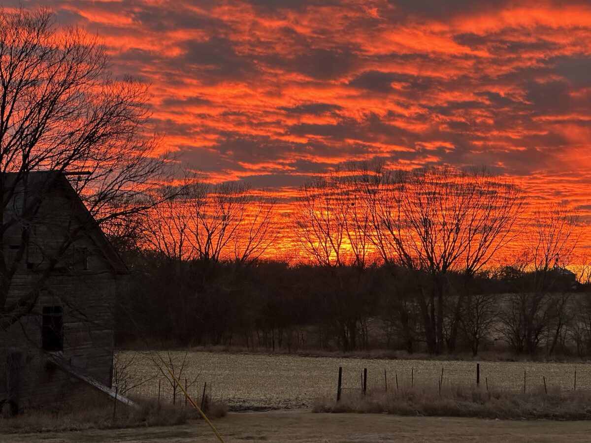 A fiery orange sky filled with clouds at sunset. In the foreground, to the left, stands the dimly lit silhouette of an old barn. To the right is an empty, harvested cornfield bordered by a tree line near the horizon.