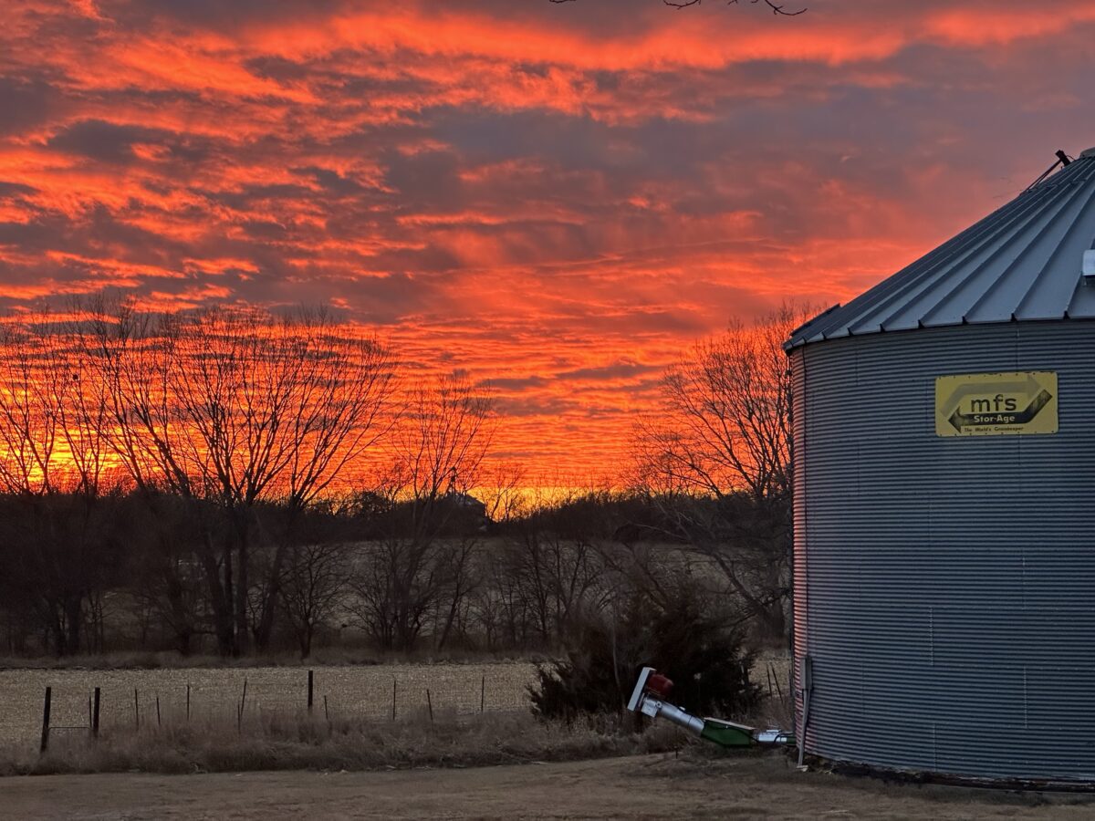 A fiery orange sunset over rolling hills of harvested cornfields, bordered near the horizon by the silhouettes of leafless trees. To the right, in the foreground, stands a silver grain silo.